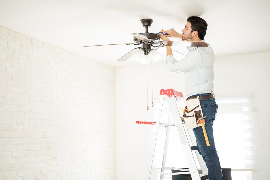 An electrician standing on a ladder installing a ceiling fan.